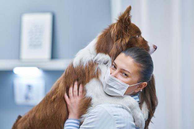 A woman wearing a face mask embraces a large brown and white dog, holding it close while smiling. The background appears to be an indoor setting, possibly a veterinary clinic or pet care facility.